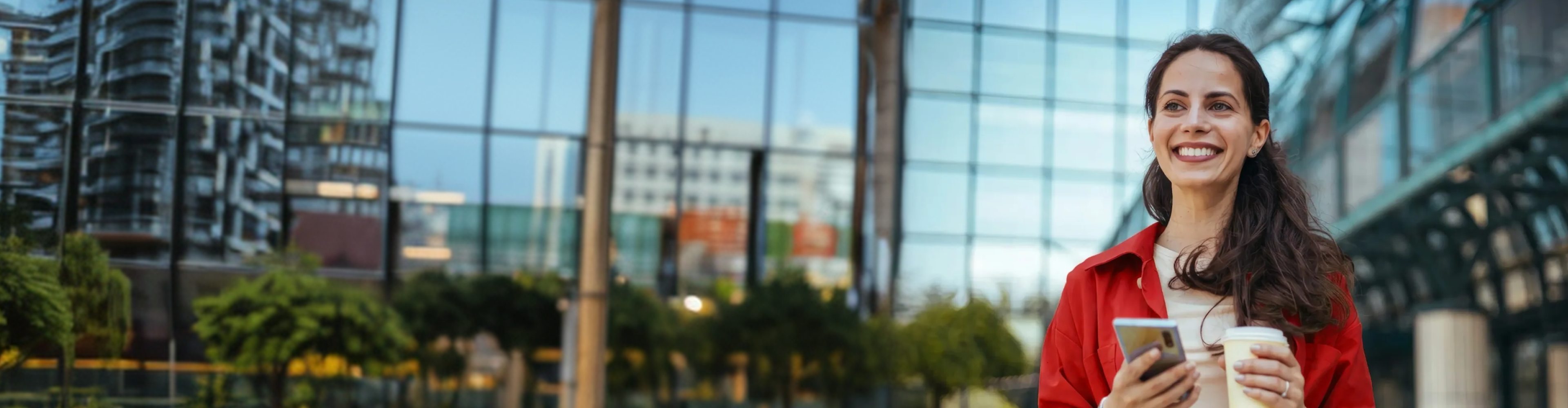 Mulher sorri com smartphone e café na mão porque tem Conta Santander Mais