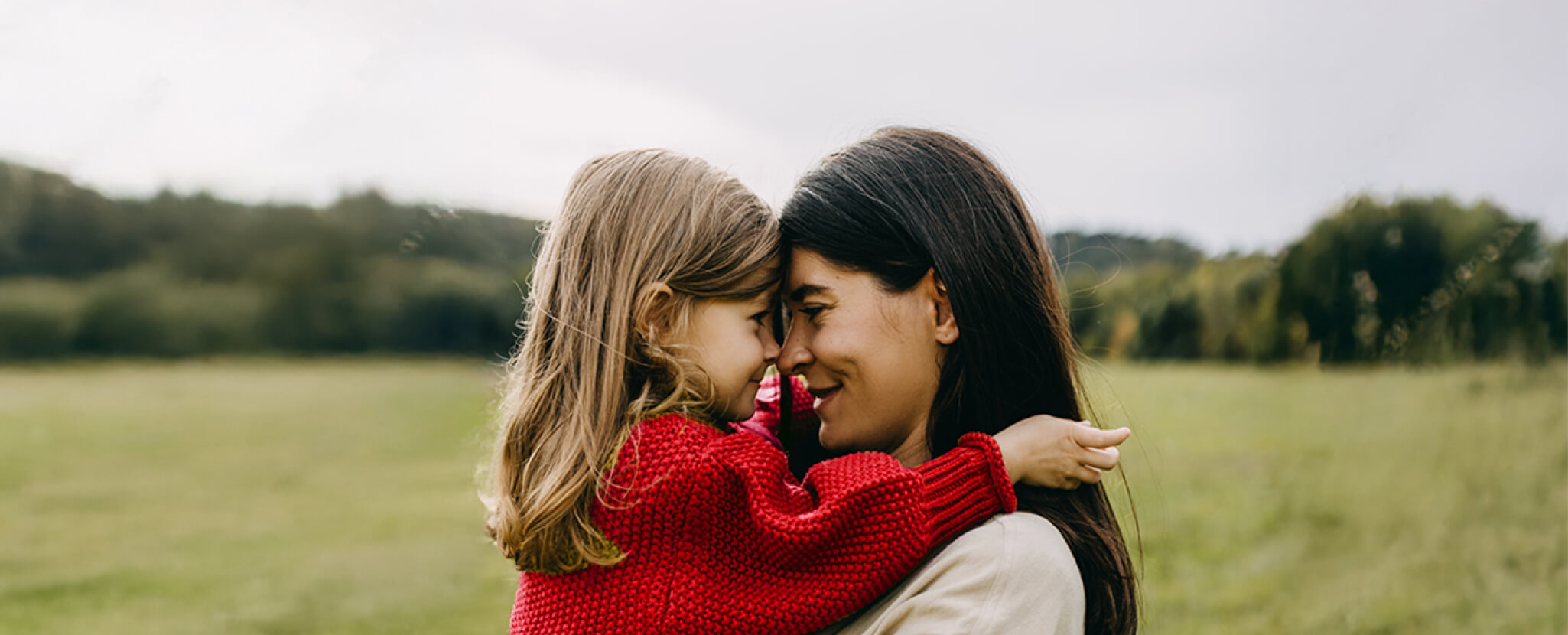 Mulher olha nos olhos da criança que tem ao colo em paisagem bucólica