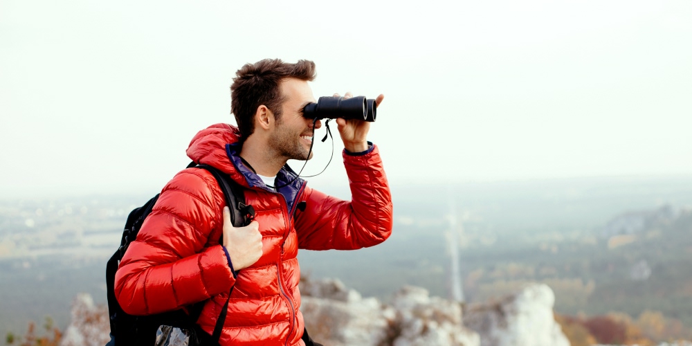 Homem de kispo e mochila às costas admira paisagem através de binóculos