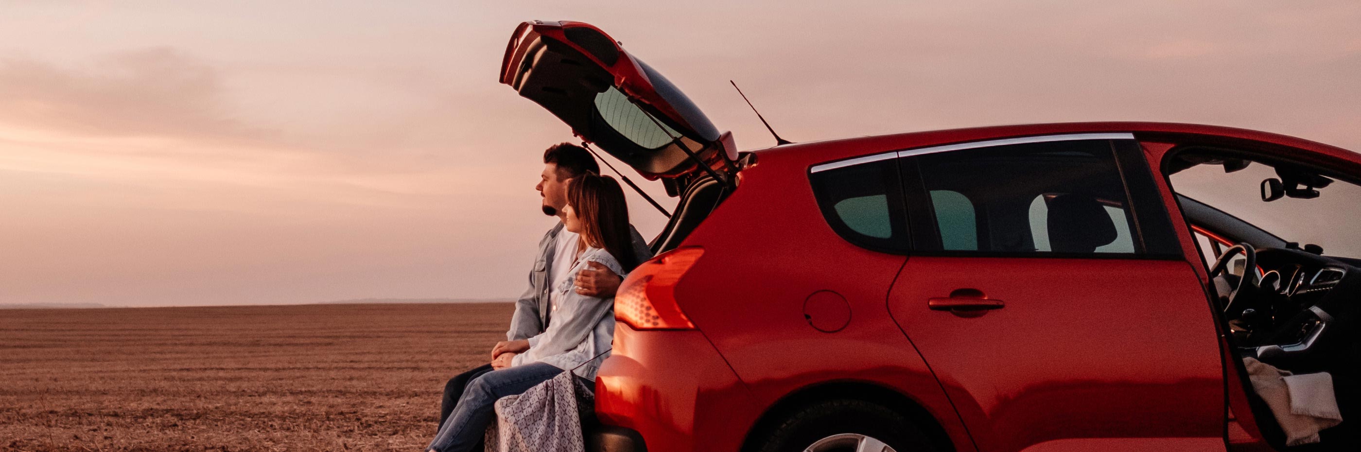 Casal de homem e mulher abraçados sentados na mala do carro contemplam pôr do sol na praia