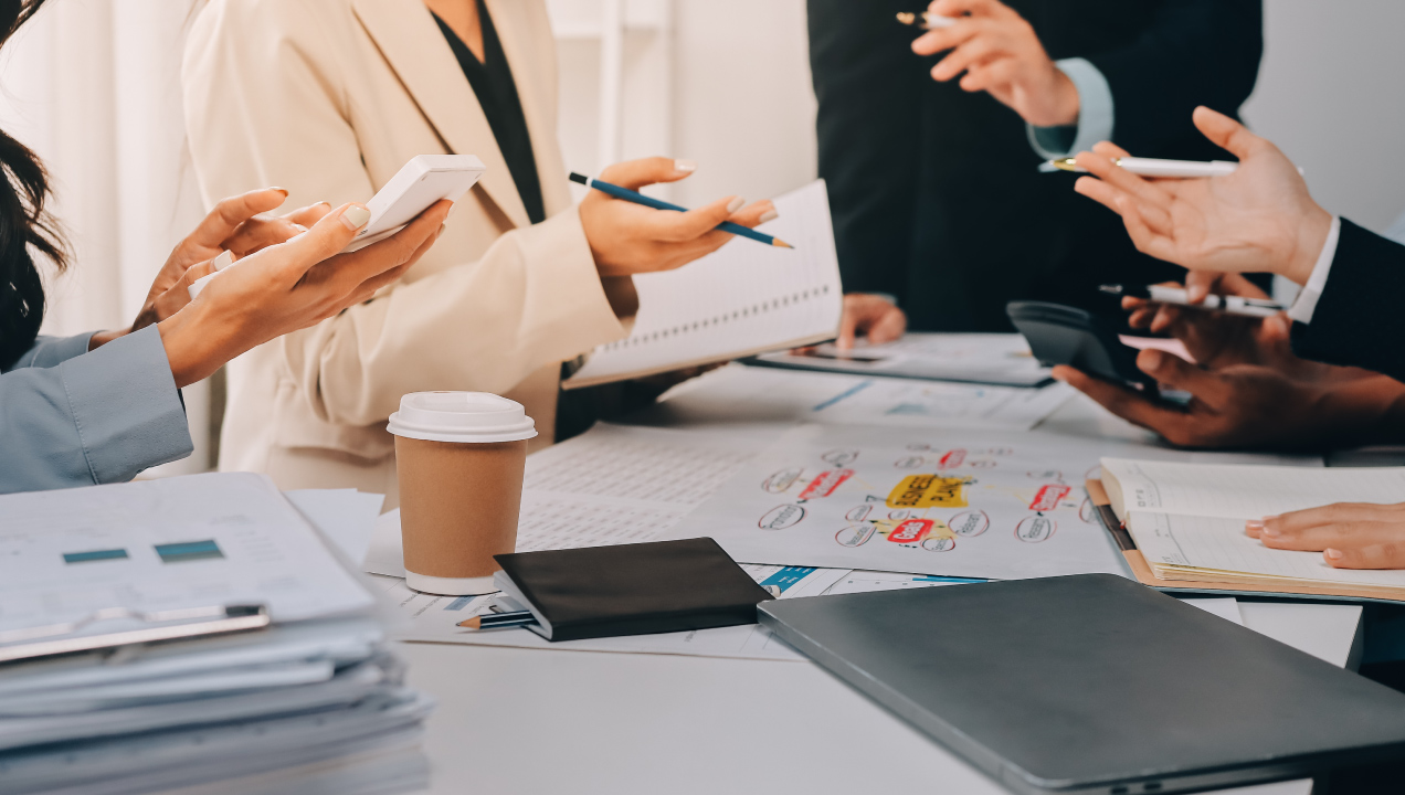 Equipa reunida à volta de uma mesa a analisar documentos e a utilizar dispositivos