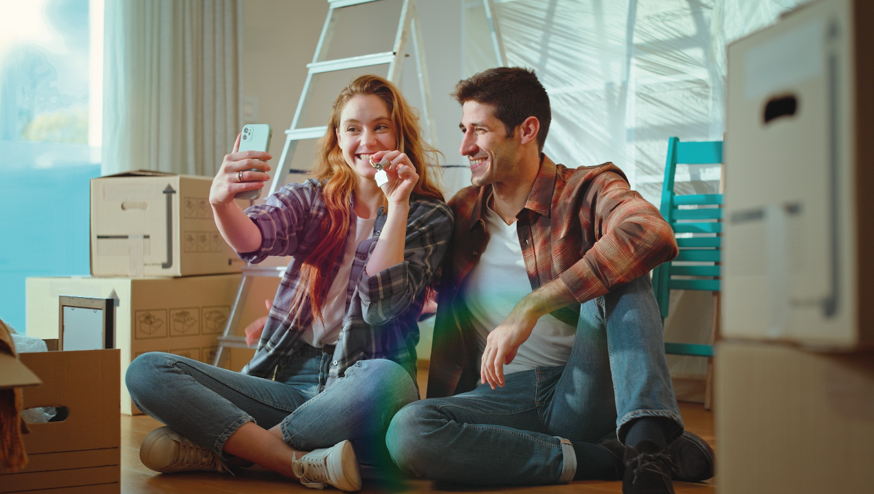 Casal jovem sentados no chão da casa felizes e a tirarem uma fotografia a segurar as chaves de casa
