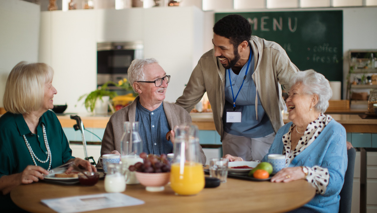 Idosos sentados a lancharem e a conversarem animados com o auxiliar