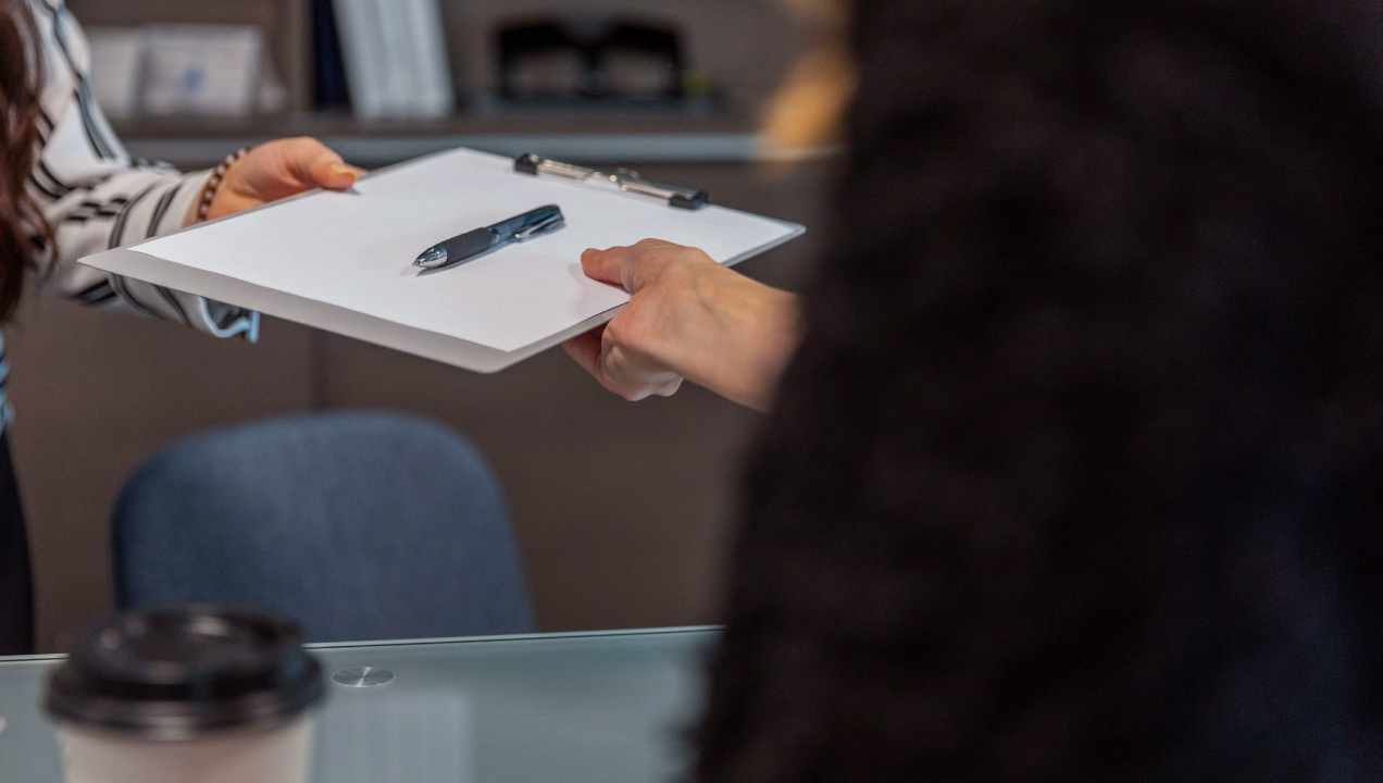 Duas pessoas de pé a segurarem uma prancheta com um documento e uma caneta, num contexto de reunião ou assinatura de documentos