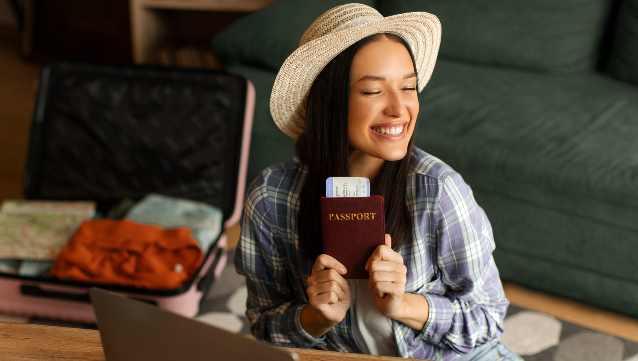 Mulher jovem sorridente a segurar um passaporte e cartão de embarque ao lado de uma mala de viagem