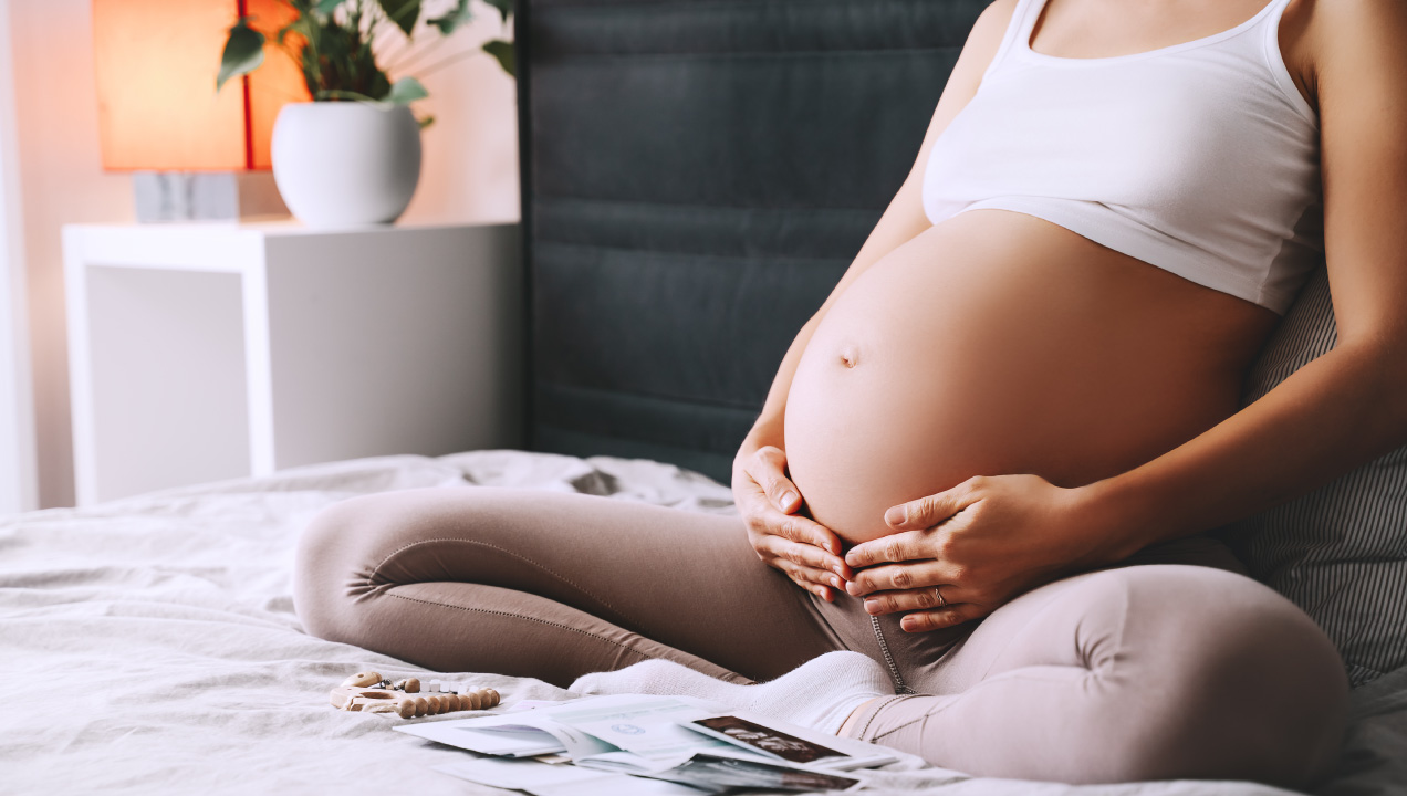 Senhora grávida sentada na cama a segurar a barriga e com documentos à frente