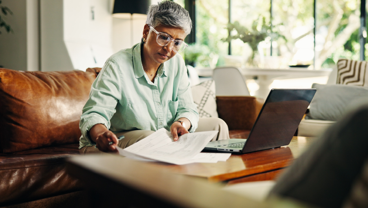 Senhora sentada no sofá de frente ao computador a mexer em documentos