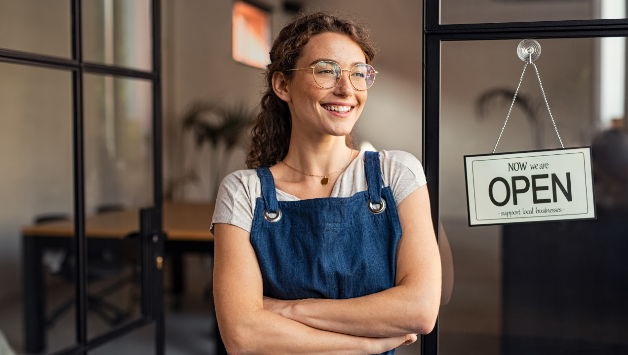 Empresária sorridente à porta do seu estabelecimento com placa a dizer aberto
