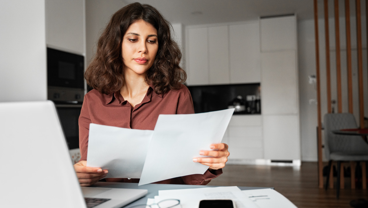 Senhora jovem a analisar documentos sentada e com o computador à frente