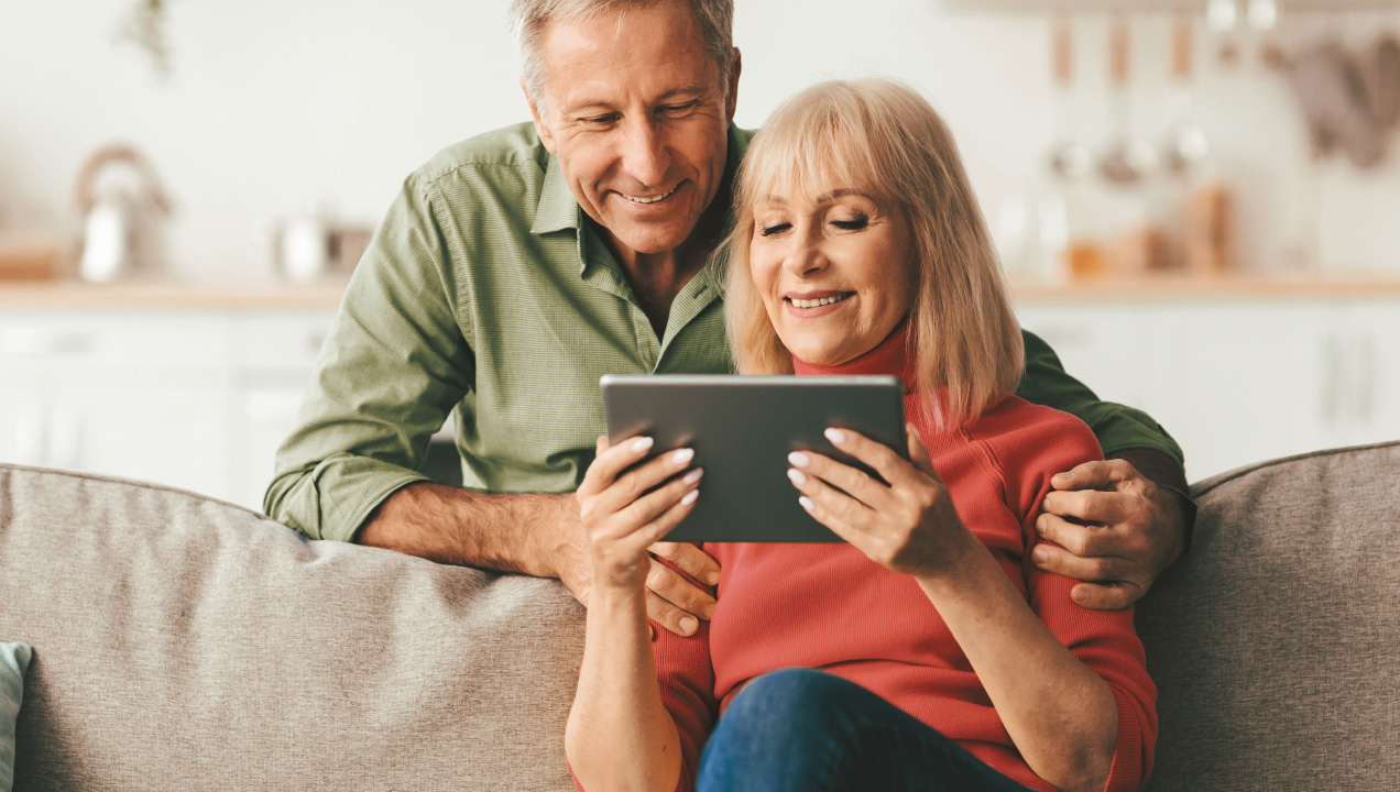 Senhora a mexer no tablet enquanto o marido lhe dá um abraço 