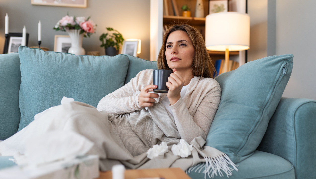 Senhora doente deitada no sofá a beber um chá e com medicação e lenços à volta dela