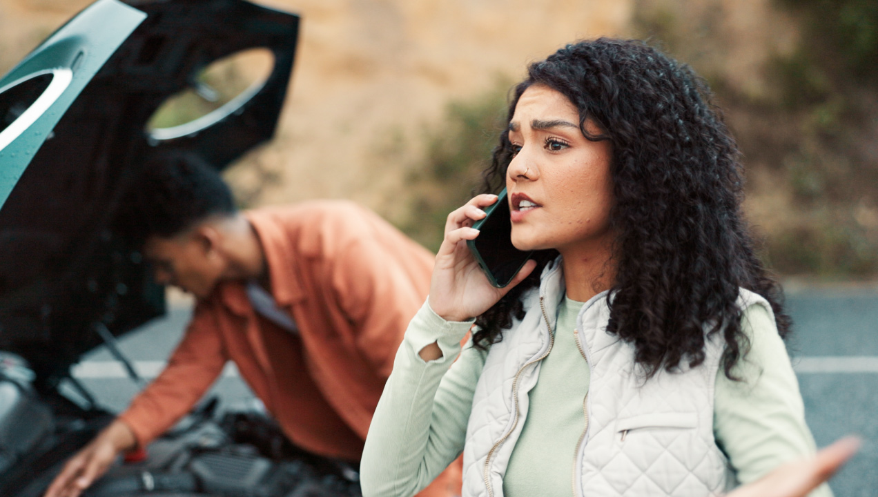 Mulher a falar ao telemóvel ao lado de um carro avariado enquanto um senhor verifica o veículo