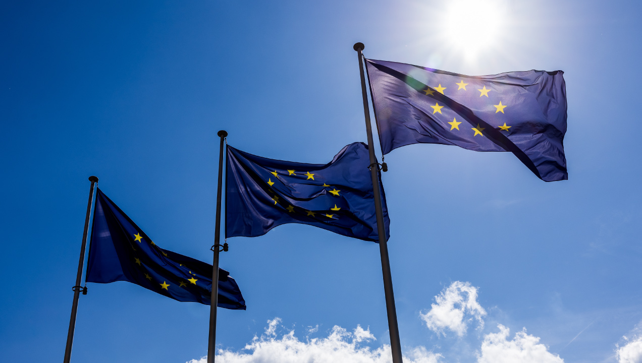 Três bandeiras da União Europeia asteadas contra um céu azul com nuvens brancas