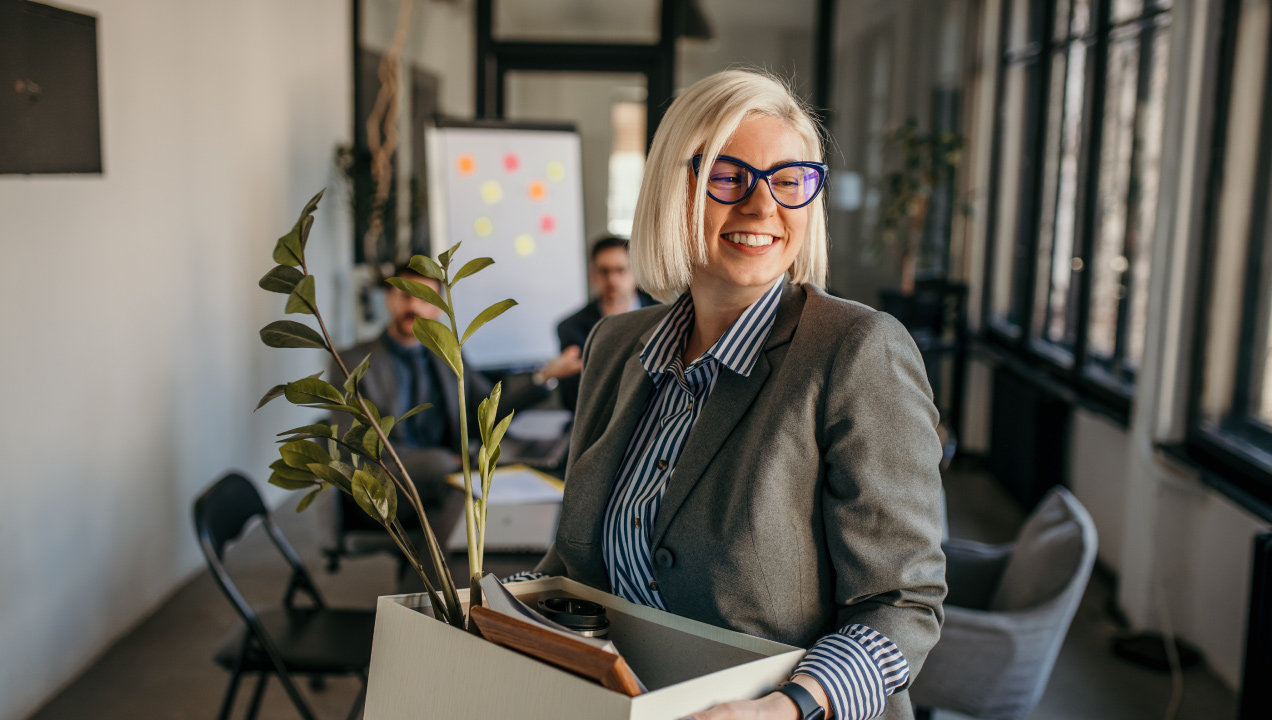 Senhora feliz a sair do escritório a segurar uma caixa com os seus artigos 