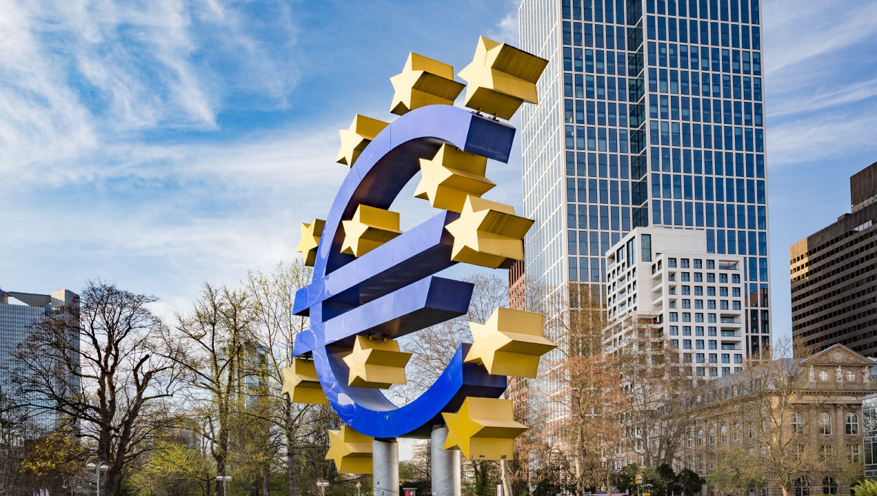 Símbolo do euro rodeado das estrelas da União Europeia, junto a edificios financeiros