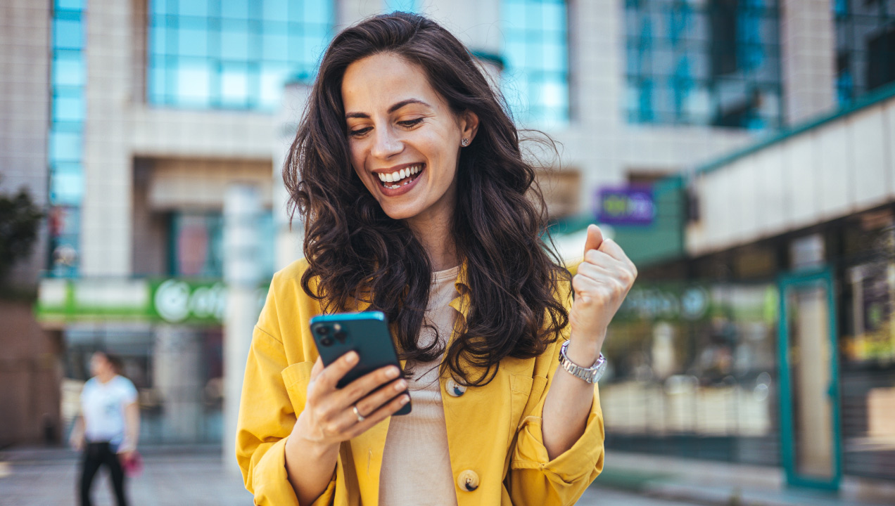 Senhora está a segurar o seu telemóvel, com a mão levantada a celebrar. Estás no exterior muito feliz e a celebrar a sua conquista.