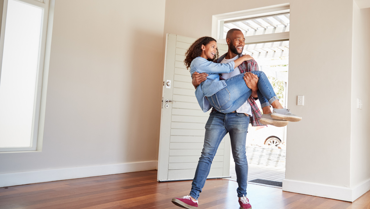 Casal feliz a entrar dentro de uma casa, senhor a segurar a senhora ao colo