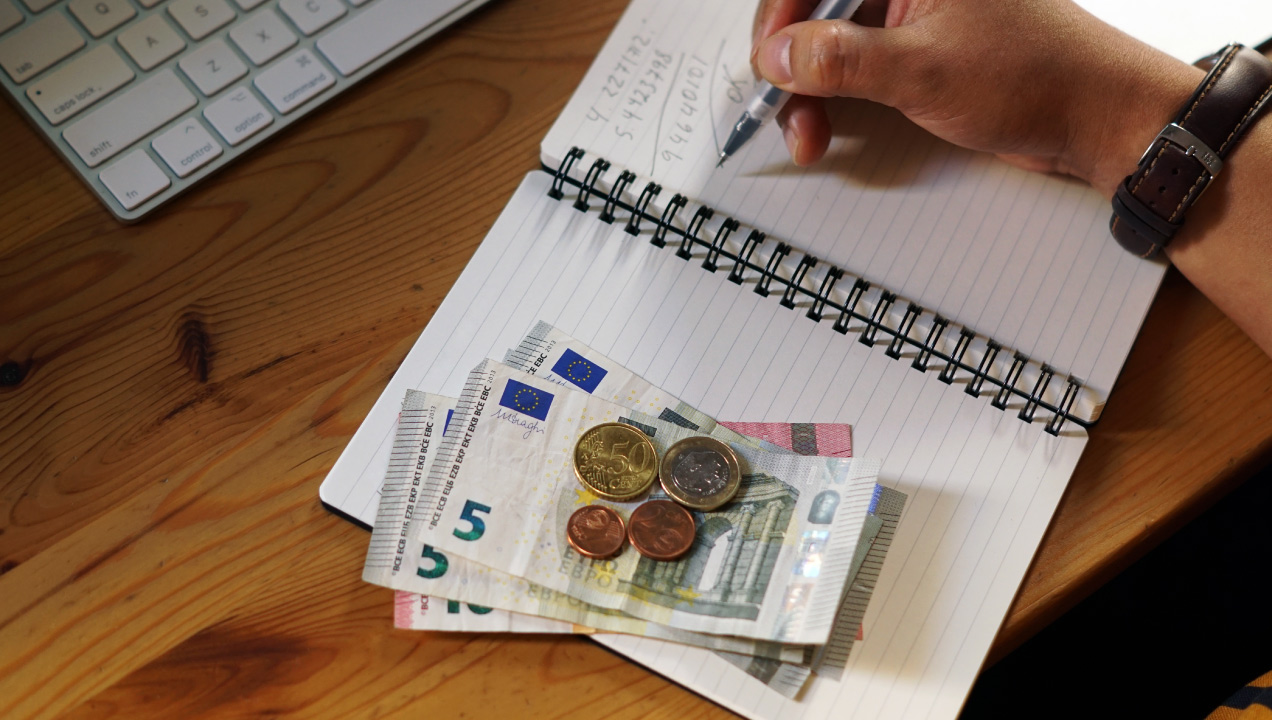 Mão a escrever um caderno e a fazer contas e ao lado do caderno algumas notas e dinheiro