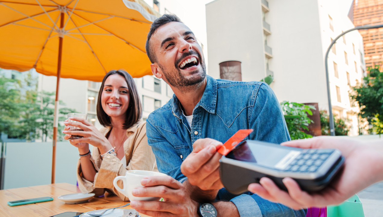 Casal sentado no exterior de um café felizes. O homem está a pagar com o cartão de crédito contactless.