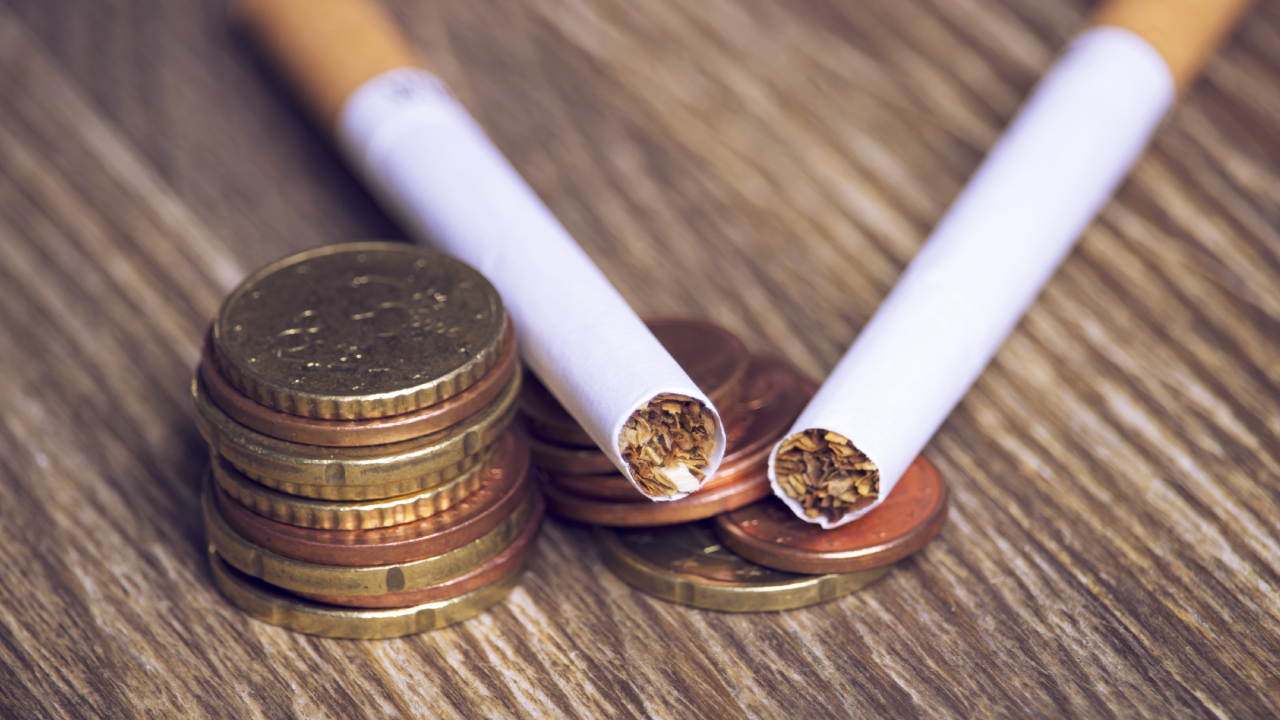 Fotografia de dois cigarros e de uma pilha de moedas, para ilustrar o imposto a pagar pelo consumo de, neste caso, tabaco.