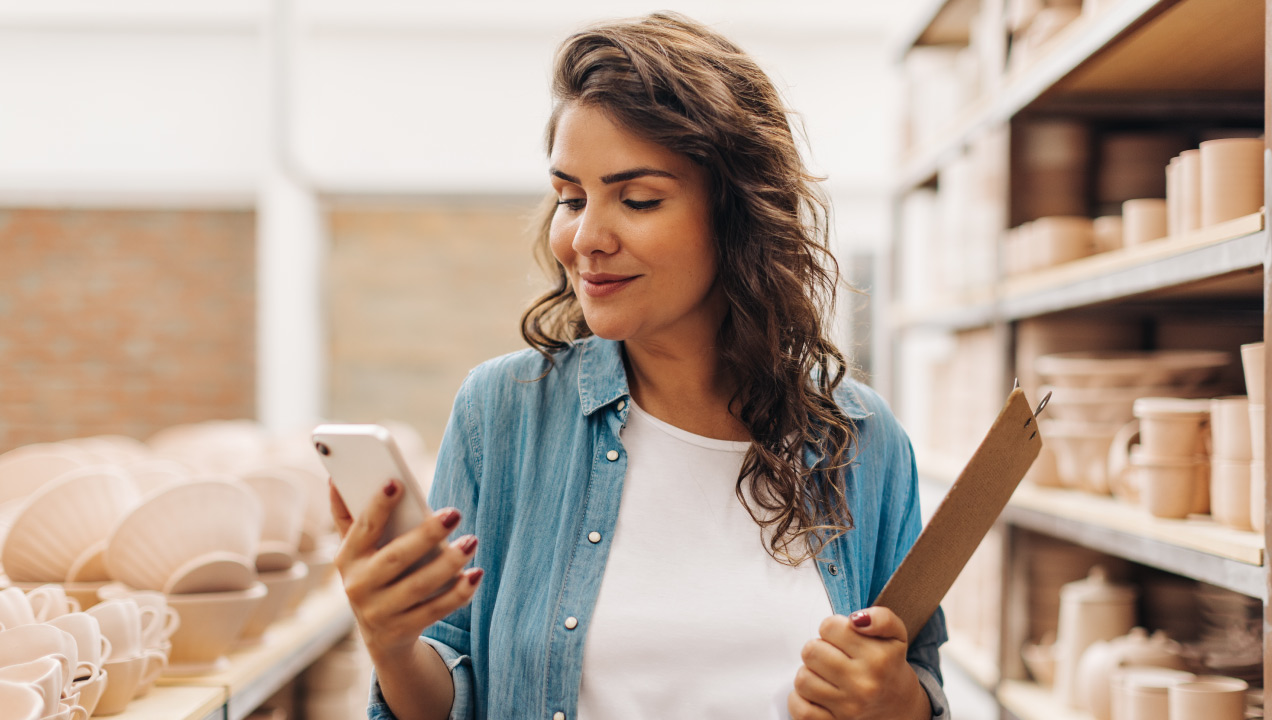 Senhora a segurar o telemóvel, sorrindo, numa loja rodeada de prateleiras com peças de cerâmica 