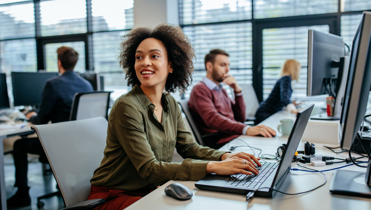 Senhora sentada numa secretária de escritório e mexer no computador, feliz e em ambiente corporativo