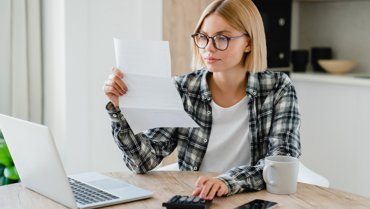 Senhora sentada numa mesa em frente ao computador, a fazer contas na calculadora enquando analisa uma folha