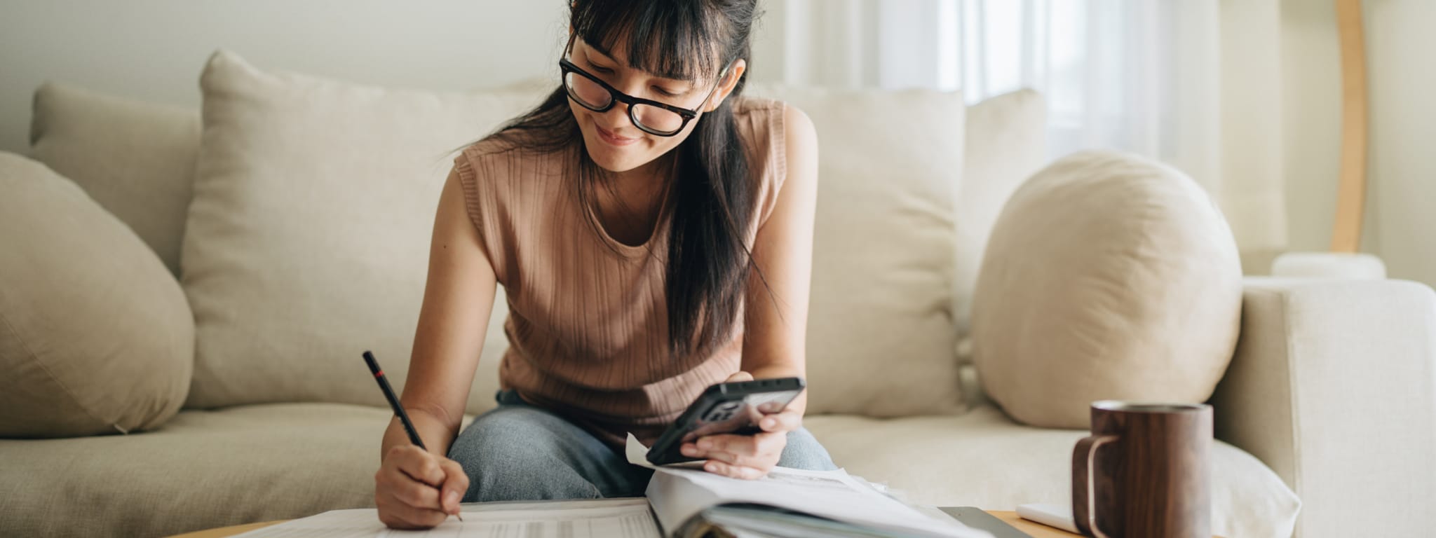 senhora sentada no sofá com o telemóvel na mão e a fazer calculos à mão