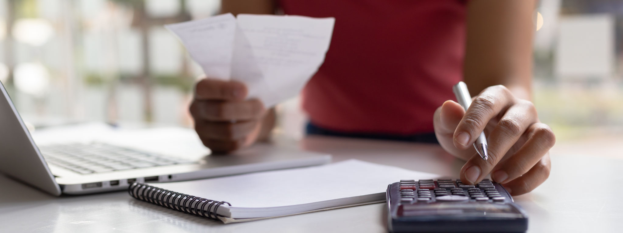 Pessoa a fazer cálculos numa calculadora, com um bloco de notas e um computador portátil ao lado.