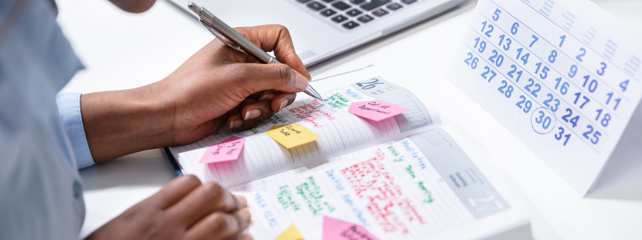  alguém a tomar notas numa agenda com post-its junto a calendário de secretária