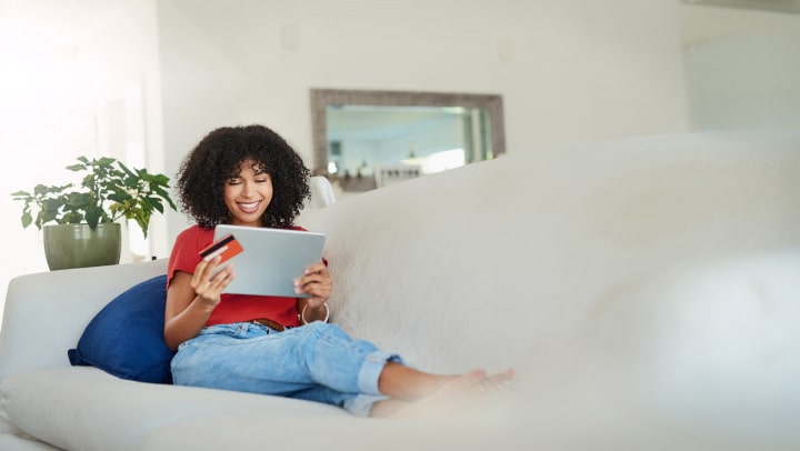 senhora com um tablet e um cartão bancário na mão sentada no sofá
