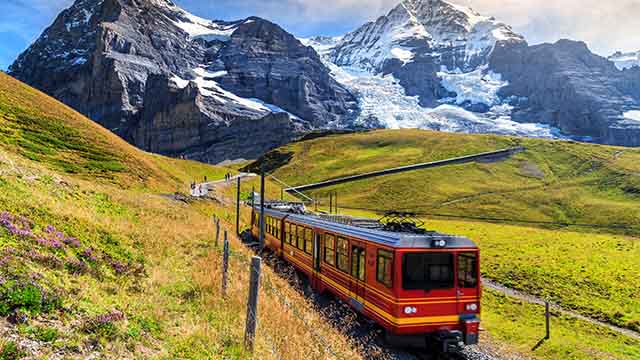 imagem de comboio a viajar por entre prados e com montanhas com neve atrás