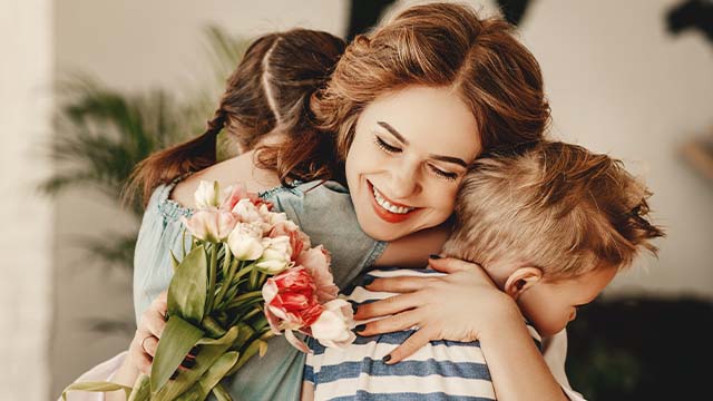 mãe a abraçar os dois filhos com um ramo de flores nas mãos
